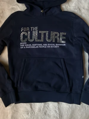 Mörkblå hoodie med text och strass - Mörkblå hoodie med huva och magficka. På bröstet står det 'FOR THE CULTURE' där 'CULTURE' är dekorerat med strassdetaljer. Under texten finns en definition i vitt tryck. Hoodien har ribbade muddar och avslappnad passform.