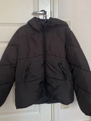 Svart pufferjacka med huva - Säljer en svart pufferjacka med huva och dragkedja framtill. Jackan har två snedställda fickor med knappstängning och en oversized passform. Perfekt för kalla dagar och riktigt skön att bära. Inte använd så mycket så ganska bra skick