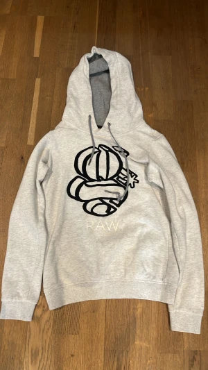 Grå hoodie från G-Star RAW XS - Snygg grå hoodie från G-Star RAW i slim fit-modell. Stor svart grafisk print på bröstet och texten RAW under. Bre skick