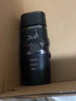 Lattafa Asad Elixir parfym - Lattafa Asad Elixir parfym i en snygg svart flaska med orientaliska detaljer och silverfärgad text. Flaskan har en robust design och lock i svart plast. Parfymen har en ljusgul vätska och ger ett exklusivt intryck.
