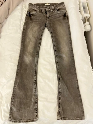 Grå bootcut jeans med kontrastsömmar - Säljer ett par grå bootcut jeans med snygga kontrastsömmar och detaljerade bakfickor. Jeansen har låg midja och klassisk femficksdesign. Perfekta för dig som gillar en avslappnad men trendig stil. Materialet är denim i bomull.