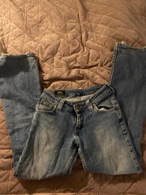 Blå slitna bootcut jeans från Lee - Säljer ett par blå bootcut jeans från Lee med slitningar på knäna och råa kanter vid bensluten. Klassisk femficksmodell i denim med en avslappnad passform och vintagekänsla. Perfekta för en cool och avslappnad stil.