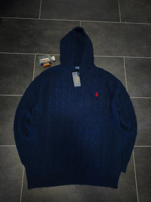 Unik hoodie från Polo Ralph Lauren - Snygg mörkblå hoodie från Polo Ralph Lauren med kabelstickat mönster och röd broderad logga på bröstet. Tröjan har huva och ribbade muddar vid ärmslut och nederkant. Perfekt för en stilren och avslappnad look. 