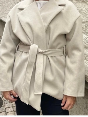 Beige kappa med knytskärp - Stilren beige kappa med bred krage och knytskärp i midjan. Kappan har en rak passform och är tillverkad i ett mjukt, ull-liknande material. Perfekt för lager-på-lager och enkel att matcha med olika outfits. Lite lite kort i armarna men inget man direkt tänker på enligt mig. 