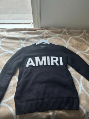 Svart stickad tröja från Amiri - Snygg svart stickad tröja från Amiri med stor vit logga över bröstet. Tröjan har rund halsringning, långa ärmar och ribbstickade muddar vid ärmslut och nederkant. Perfekt för dig som gillar streetwear och vill ha något stilrent.