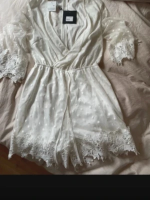 Vitt nattlinne med spets från Hunkemöller - Säljer ett vitt nattlinne från Hunkemöller med vackra spetsdetaljer på ärmar och nederkant. Nattlinnet har prickigt mönster, omlott-design och knyts i midjan. Materialet är lätt och transparent, perfekt för en mysig kväll hemma.