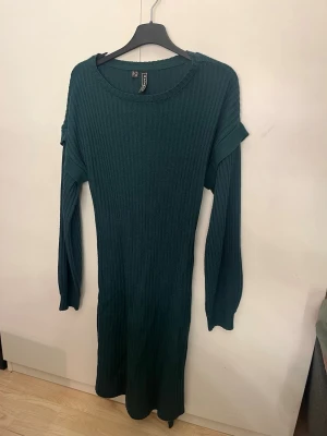 Mörkgrön ribbad stickad klänning - Mysig mörkgrön stickad klänning från märket rainbow med ribbad struktur. Klänningen har långa ärmar och en volangdetalj vid axlarna som ger en extra touch. Perfekt för kyligare dagar och enkel att styla med boots eller sneakers.