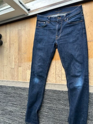 Mörkblå jeans från Nudie Jeans - Säljer ett par mörkblå jeans från Nudie Jeans med klassisk femficksmodell och kontrastsömmar. Jeansen har raka ben och Nudie Jeans ikoniska broderi på bakfickorna. Perfekta för en avslappnad och stilren look.