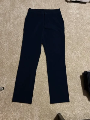 Mörkblå byxor från Under Armour - Snygga mörkblå byxor från Under Armour med rak passform och klassisk design. Byxorna har knapp och dragkedja framtill samt diskreta fickor. Perfekta för dig som gillar stilrena och bekväma jeans med sportig känsla.