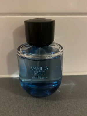 Zara Vanilla Melt parfym blå - Vanilla Melt från Zara Bloom Collection är en parfym i en rund, blå glasflaska med svart lock. Flaskan har en modern och stilren design med tydlig text på framsidan. Doften är inspirerad av vanilj och passar dig som gillar söta toner.