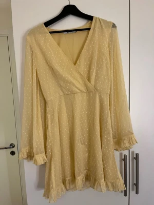 Ljusgul prickig omlottklänning - Söt ljusgul kortklänning med omlottdesign. Klänningen har långa ärmar med volangdetaljer vid ärmslut och nederkant. V-ringad och i ett lätt, luftigt material som passar perfekt till sommaren .