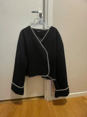 Svart omlottkofta med vita sömmar - Syrorlek L men sitter som S-M. Stilren svart omlottkofta med vita kontrastsömmar längs kanterna och ärmsluten. Koftan har en croppad passform och raka, långa ärmar. Perfekt för att lyfta en enkel outfit med en unik twist. Båda knapparna fungerar ej tyvärr men går att använda som en kofta som man slänger över liksom, ena knappen är helt av 