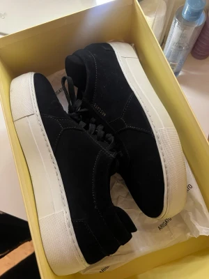 Svarta sneakers från Axel Arigato - Snygga svarta sneakers från Axel Arigato med ovandel i mocka och klassisk vit tjock sula. Diskret guldfärgad logga på sidan och svarta skosnören. Perfekta för dig som gillar stilrena och trendiga skor.