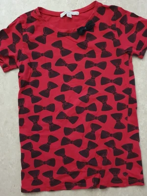 T-shirt - Cool röd t-shirt med svarta rosetter över hela tröjan och en extra tygrosett vid halsringningen. T-shirten har rund hals och korta ärmar, perfekt för dig som vill sticka ut lite extra. Tillverkad i mjuk bomull som känns skön mot huden.