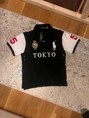 Svart och vit pikétröja Polo Tokyo - Snygg svart pikétröja från Polo Ralph Lauren med vita ärmar och stora tryck. Framsidan har TOKYO-text, logga och emblem, medan baksidan har en stor röd femma. Klassisk krage och korta ärmar, perfekt till en sportig och trendig stil.