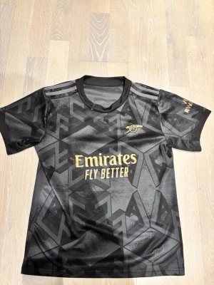 Arsenal svart matchtröja Saka 7 - Snygg svart Arsenal matchtröja från Adidas med guldiga detaljer och trycket 'Emirates Fly Better' på bröstet. På ryggen står det 'Saka' och nummer 7 i vitt. Tröjan har korta ärmar, rund hals och ett grafiskt mönster över hela tyget. Perfekt för fotbollsfans!