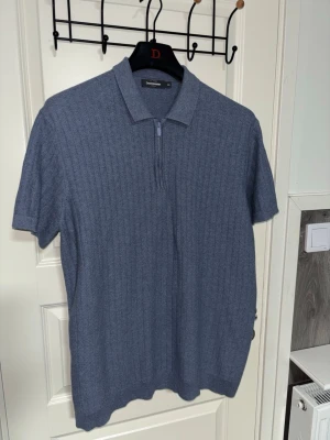 Blå stickad pikétröja Dressmann - Snygg blå stickad pikétröja från Dressmann med kort ärm och dragkedja vid halsen. Tröjan har diskret ribbat mönster och klassisk krage, perfekt för en stilren och avslappnad look. Materialet känns mjukt och bekvämt.