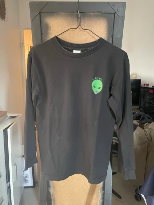 Svart långärmad topp från RIPNDIP med alien-tryck - Cool svart långärmad topp från ripndip med grönt alien-tryck och texten 'We Out Here' på bröstet och ryggen. Tryck i grönt även på ena ärmen. Perfekt för dig som gillar streetwear och vill sticka ut lite extra.