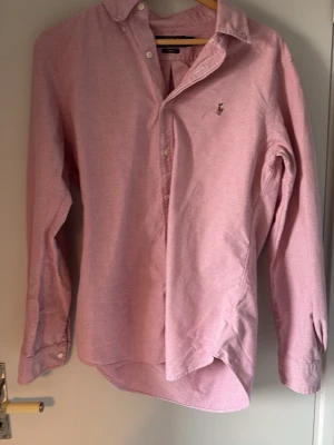Rosa skjorta från Ralph Lauren - Klassisk rosa skjorta från Ralph Lauren med button-down krage och broderad logga på bröstet. Skjortan har lång ärm, vita knappar och är tillverkad i mjuk bomull. Perfekt för en clean och stilren look.