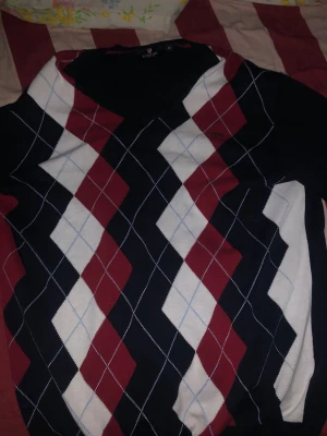 Stickad tröja med argylemönster från U.S polo assn  - Snygg stickad tröja från us. Polo assn med klassiskt argylemönster i svart, vinrött och vitt. Tröjan har V-ringning och långa ärmar. Perfekt för dig som gillar preppy stil och vill ha något som sticker ut lite extra.