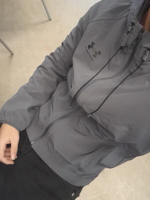 Grå vindjacka från Under Armour - Snygg grå vindjacka från Under Armour med dragkedja framtill och justerbar huva. Jackan har en sportig look med logga på bröstet och är tillverkad i ett lätt, vindtätt material. Perfekt för dig som vill ha en stilren och funktionell jacka.