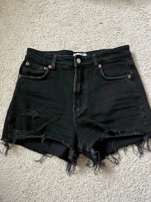 Svarta högmidjade jeansshorts - Säljer ett par svarta högmidjade jeansshorts med råa, fransiga kanter och slitna detaljer framtill. 