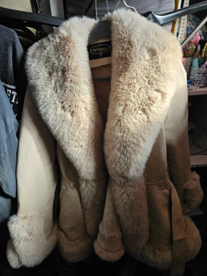 Beige fuskpäls-kappa från Profimod - Snygg beige kappa från Profimod med bred krage och muddar i mjuk fuskpäls. Kappan har markerad midja med bälte och stora fickor framtill. Perfekt för dig som vill ha en varm och trendig look med lyxig känsla. Pälsen är fluffig och kappan har en klassisk, oversized passform.