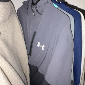 Grå vindjacka från Under Armour - Snygg grå vindjacka från Under Armour med vit logga på bröstet och dragkedja på bröstfickan. Jackan har huva och är perfekt för blåsiga dagar. Sportig look och passar dig som gillar streetwear.