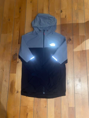 Grå och svart vindjacka från The North Face - Snygg vindjacka från The North Face i grått och svart med huva och hel dragkedja. Jackan har reflekterande detaljer på bröstet och ärmarna, perfekt för att synas i mörker. Materialet är lätt och funktionellt, idealiskt för utomhusaktiviteter. PRIS KAN SÄNKAS VID SNABB AFFÄR!!