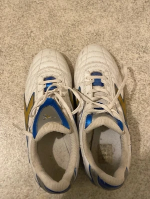 Vita sneakers med blå och gula detaljer - Säljer ett par vita sneakers från Diadora med blå och gula detaljer på sidorna och tungan. Skorna har klassisk låg profil och är tillverkade i syntetmaterial med textilfoder. Perfekta för dig som gillar sportig stil och vill sticka ut med färg.