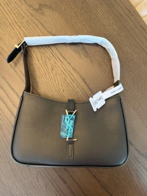 Svart axelremsväska från Saint Laurent - Snygg svart handväska från Saint Laurent i slätt läder med silver YSL-logga framtill. Väskan hare en stilren, rektangulär form och kort axelrem . Perfekt för dig som gillar klassisk och lyxig design. Dustbag ingår. Aldrig använd 