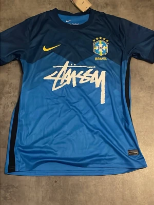 Blå Brasil Stüssy x Nike fotbollströja - Säljer en blå fotbollströja från Nike i samarbete med Stüssy och Brasil. Tröjan har mörkblå axlar, Brasil-logga med stjärnor, gul Nike-logga och stort vitt Stüssy-tryck på bröstet. Materialet är lätt och andas, perfekt för sport och träning.