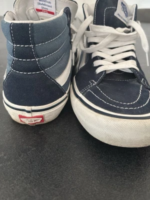 Blåa Vans Sk8-Hi Pro sneakers - Säljer ett par blåa Vans Sk8-Hi Pro sneakers med vit sula och vita detaljer. Skorna har klassisk hög siluett, vit sidostripes och grov gummisula med rutmönster undertill. Materialet är en mix av mocka och canvas, perfekta för skate eller streetwear.