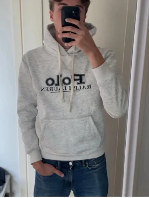 Grå hoodie från Polo Ralph Lauren - Snygg ljusgrå hoodie från Polo Ralph Lauren med stor logga framtill. Klassisk känguruficka och justerbar huva med vita snören. Mjuk bomullsblandning som är perfekt för en avslappnad stil. Toppenskick, priset ej hugget i sten vid snabb affär!