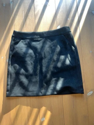 Svart mockakjol med dragkedja - Snygg svart kortkjol i mockaimitation med en cool silverfärgad dragkedja bak. Kjolen har en enkel, rak passform och är perfekt för att styla med både stickade tröjor och t-shirts. Materialet känns mjukt och ger en trendig look.