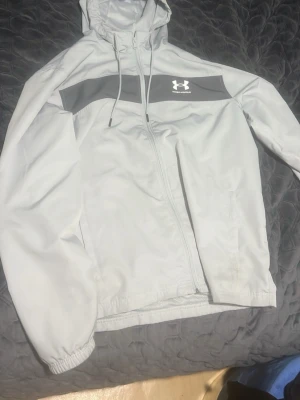 Ljusgrå vindjacka från Under Armour - Snygg ljusgrå vindjacka från Under Armour med huva och dragsko. Jackan har en bred grå panel över bröstet med logga, hel dragkedja framtill och två sidofickor. Tillverkad i lätt, vindtätt material med meshfoder för extra komfort.
