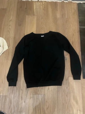 Svart basic sweatshirt - En enkel svart sweatshirt med rund hals och långa ärmar. Tröjan har ribbade muddar vid ärmslut och nederkant, perfekt för en avslappnad och stilren look. Passar bra till jeans eller joggers.