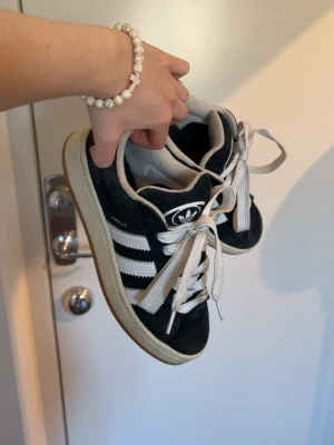 Svarta Adidas Campus sneakers - Klassiska Adidas Campus sneakers i svart mocka med vita ränder och vit sula. Skorna har vita snören och den ikoniska Adidas-loggan på plösen. Perfekta för en avslappnad streetwear-look.