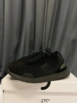 BOSS skor  - Stilrena helsvarta sneakers från BOSS med stickad ovandel och diskret logga på sidan. Skorna har en chunky sula och svarta skosnören. Perfekta för dig som gillar en clean och modern look. Materialet är textil med detaljer i syntet.