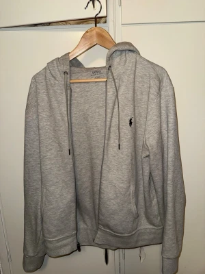 Grå hoodie från Polo Ralph Lauren - Snygg grå hoodie från Polo Ralph Lauren med dragkedja framtill och två fickor. Klassisk design med huva och justerbara snören samt den ikoniska lilla loggan broderad på bröstet. Tillverkad i mjukt bomullsmaterial som är skönt att ha på sig. Storlek M men passar S med!