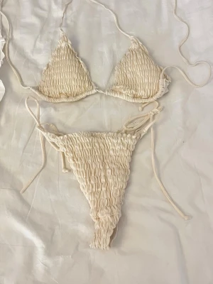 Beige bikini med smock och knyt - Trendig beige bikini med smockat tyg och knyt i sidorna. Triangeltopp med tunna band och matchande bikinitrosa med höga skärningar. Perfekt för strandhäng och soliga dagar.