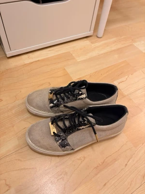 Beige sneakers med ormmönster från Polo Ralph lauren - Snygga beige sneakers från Polo Ralph Lauren med ormmönstrade detaljer och svarta skosnören. Skorna har guldfärgade metalldetaljer vid snörningen och en vit sula. Tillverkade i canvas med coola kontraster och streetkänsla. Dem är lite smutsiga men det går nog att tvätta bort