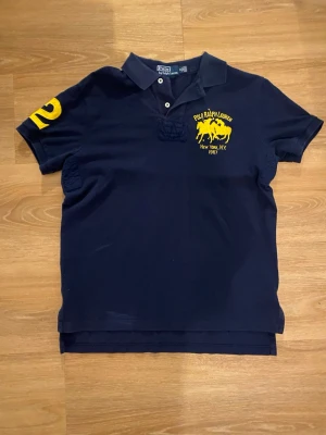 Marinblå pikétröja Polo Ralph Lauren - Klassisk marinblå pikétröja från Polo Ralph Lauren med gul logga och text på bröstet samt siffran 2 på ärmen. Tröjan har krage, knappar vid halsen och är kortärmad. Perfekt för en sportig och stilren look.
