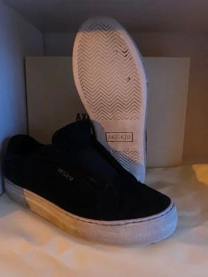 Svarta sneakers från Axel Arigato passar 39! - Stilrena svarta sneakers från Axel Arigato med vit, tjock sula och diskret logga på sidan. Ovandelen är i mocka och designen är minimalistisk utan snörning. Perfekta för dig som gillar clean och modern stil.