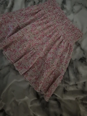 Blommig volangkjol i rosa och vit - Supersöt kort kjol med blommigt mönster i rosa och vitt. Kjolen har bred smock i midjan och volangdetaljer som ger en luftig och romantisk känsla. Perfekt för varma dagar och enkel att matcha med en enkel topp.