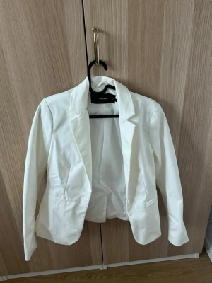 Vit kavaj från Vero Moda - Stilren vit kavaj från Vero Moda med klassisk krage och två fickor framtill. Kavajen har en figurnära passform och är tillverkad i ett mjukt tyg som ger en snygg siluett. Perfekt att matcha med jeans eller kjol för en trendig look.