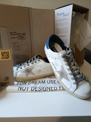 Golden Goose vita sneakers med stjärna - Golden Goose sneakers i vitt läder med grå mockastjärna på sidan, grå skosnören och blå detalj på hälen. Klassisk låg modell med slitna detaljer för en cool och avslappnad look. Perfekta för dig som gillar streetstyle och italiensk design.