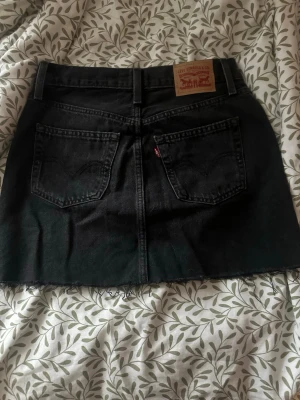 Svart jeanskjol från Levi's - Snygg svart jeanskjol från Levi's med rå kant nertill och klassiska fem fickor. Kjolen har knappgylf framtill och Levi's ikoniska läderpatch bak i midjan. Perfekt till vardags för en cool och avslappnad look. För liten för mig då ja ha M igentligen skulle säga S o xs i storleken