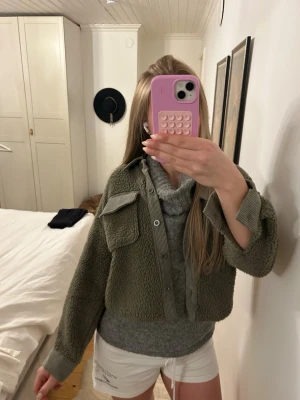 Olivgrön teddy cardiganjacka - Mysig olivgrön teddy cardiganjacka med stora bröstfickor och knappstängning framtill. Jackan har en croppad passform och ribbade muddar vid ärmsluten. Perfekt för lager på lager och ger en avslappnad streetwear-look.