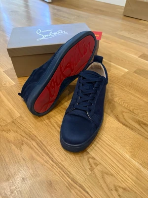 Marinblå Louboutin - Snygga marinblå sneakers från Christian Louboutin i mocka med klassisk röd sula och logotyp. Skorna har rund tå, svarta skosnören och låg profil. Perfekta för dig som vill sticka ut med exklusiv design och ikoniska detaljer.